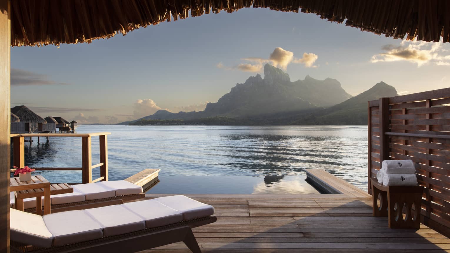 A view from a wooden Water Bungalow with white cushioned lounge chairs overlooking the ocean and mountains in the background at Four Seasons Bora Bora