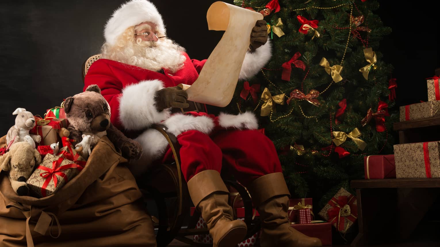 Santa Claus seated in large rocker holding a scroll, flanked by decorated Christmas tree with gifts and overflowing gift bag