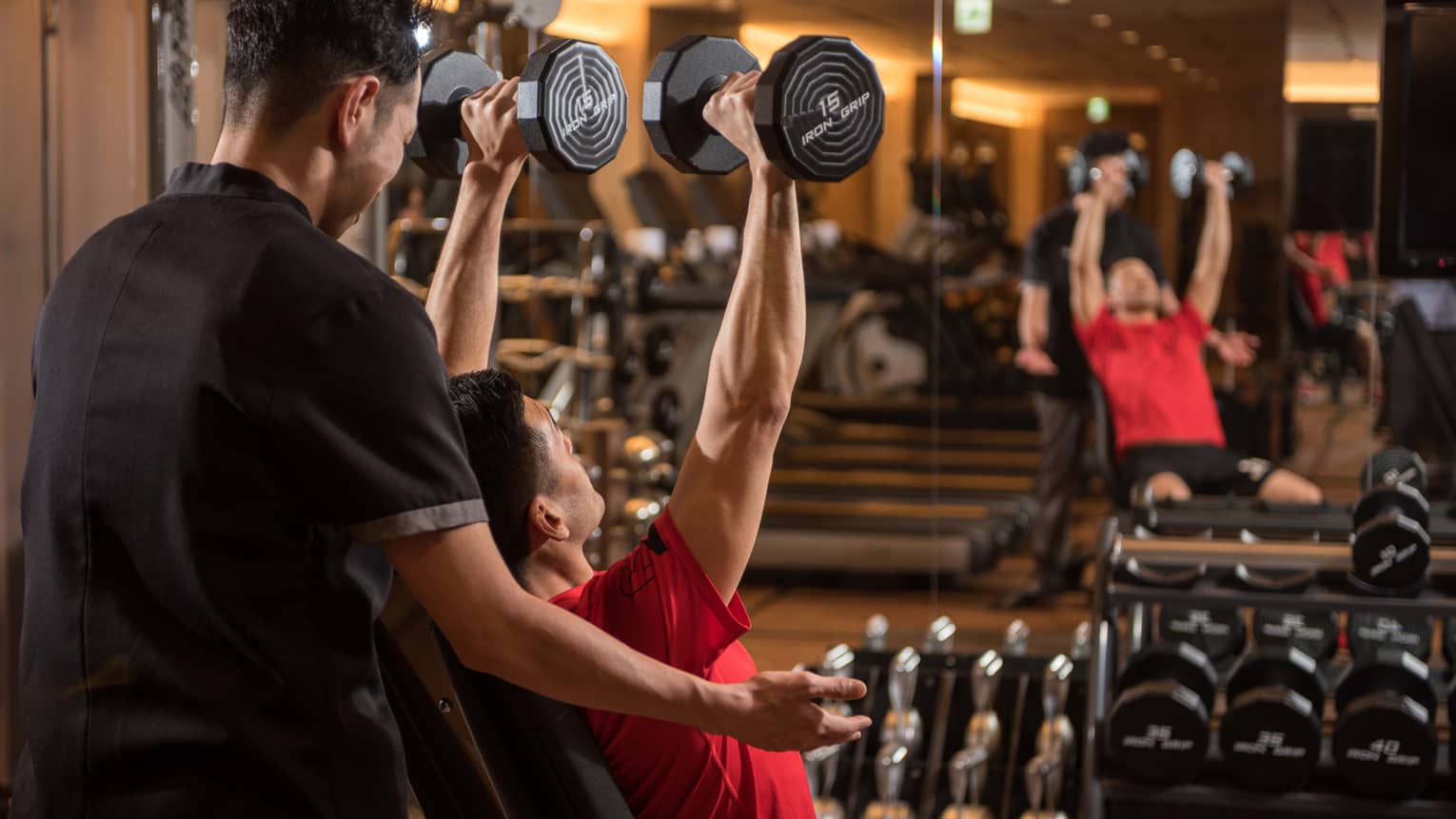 Man in chair in front of mirror lifts two 15 lb. weights above his head while personal trainer assists