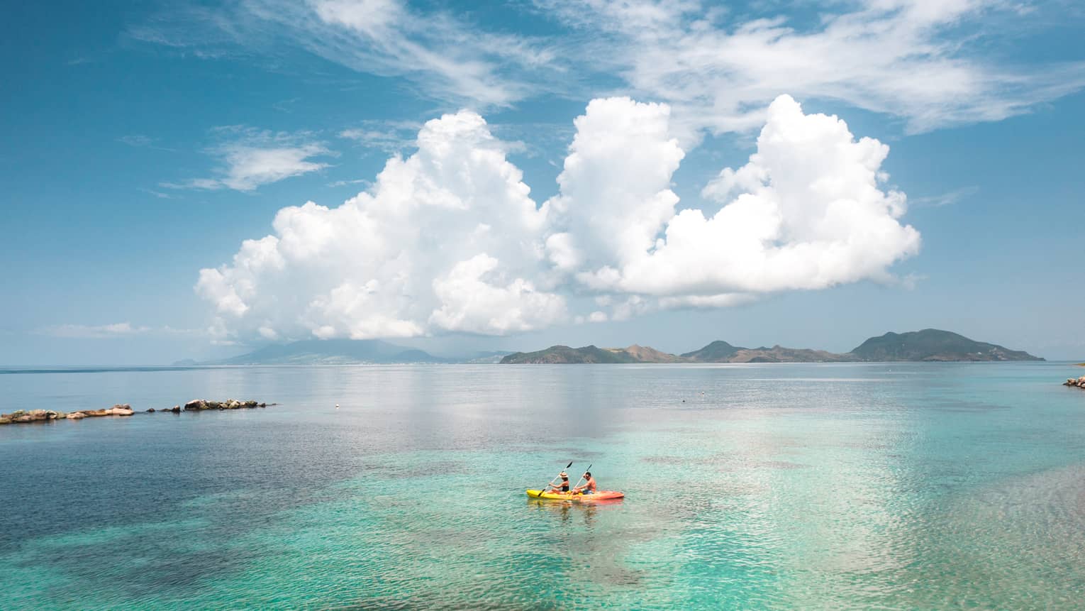 People in a yellow kayak in clear ocean water with large white clouds in the sky above them.