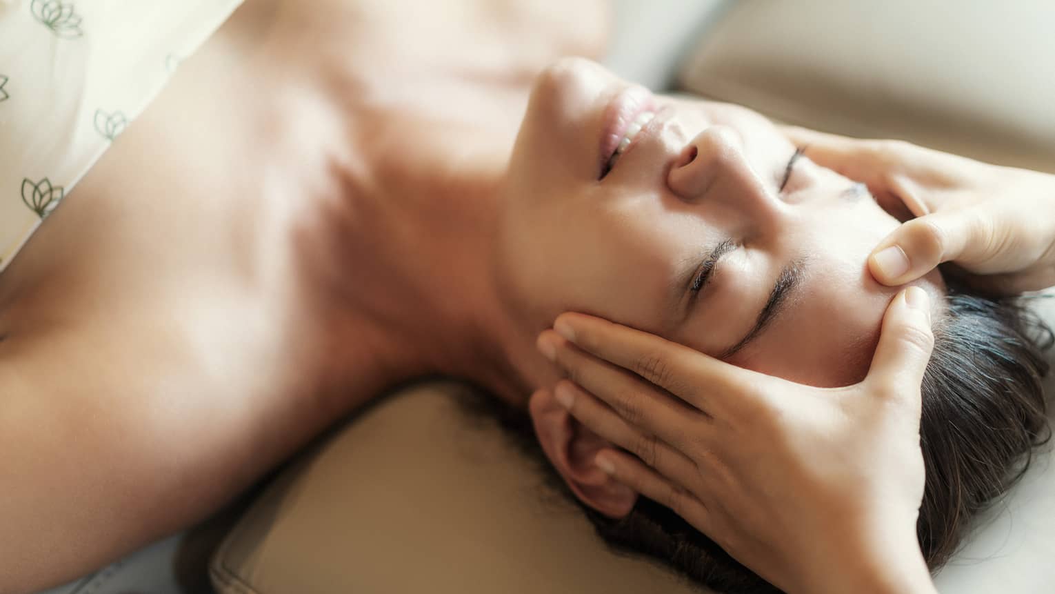 Spa facial, two hands rest on woman's forehead as she closes her eyes, lays under sheet on treatment table