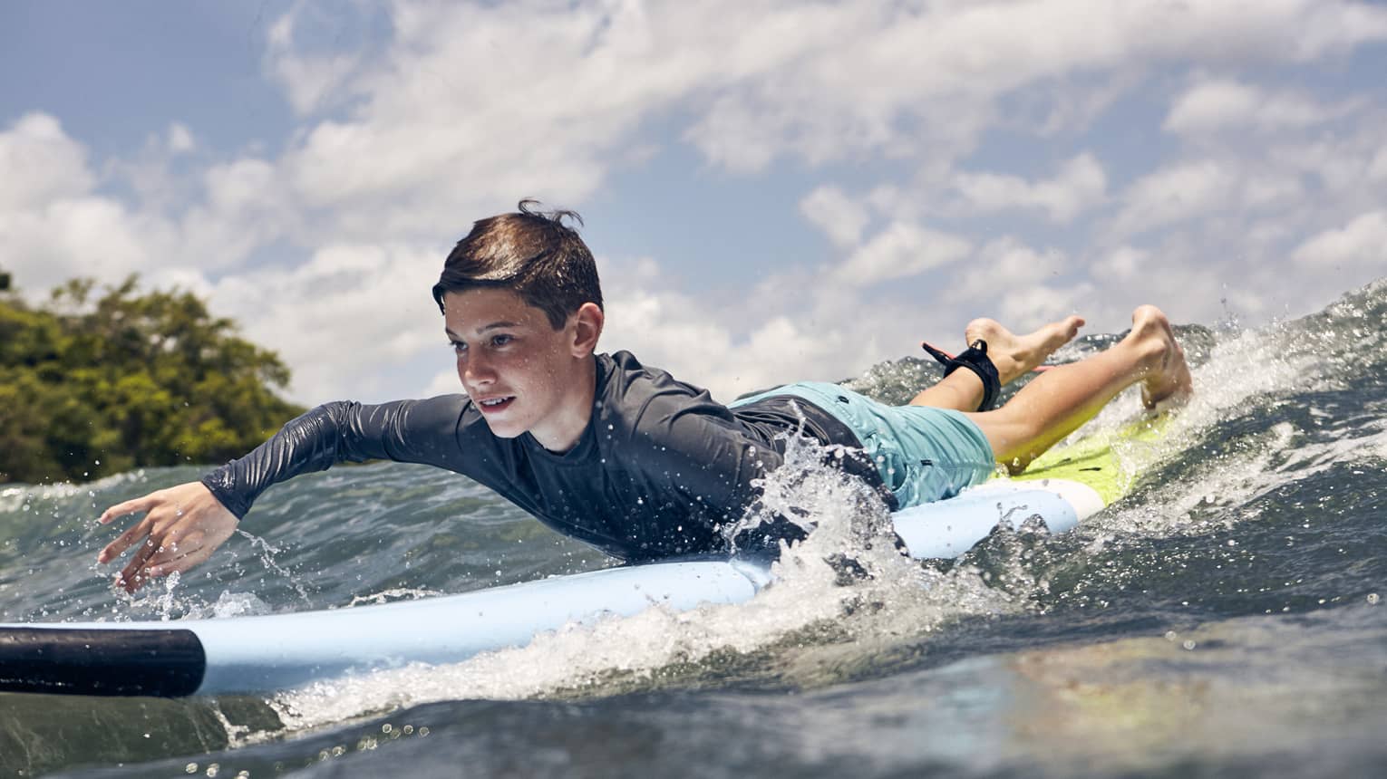 Close-up of a child paddling a surfboard over a wave in the glistening sea, water splashing around the sides of the board.