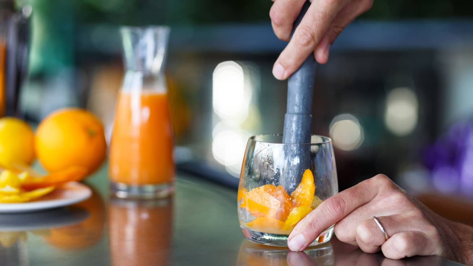 One hand steadies a clear glass while the other muddles orange slices; in the background, whole oranges, peels and a carafe.
