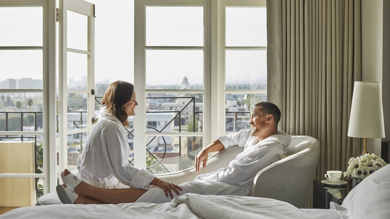 Smiling man and woman wearing bathrobes, lounging in front of sunny hotel room windows