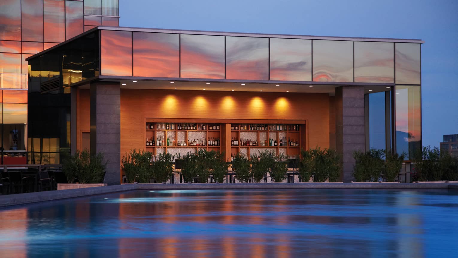 Illuminated swimming pool, wood bar and liquor displays under glass building exterior at sunset