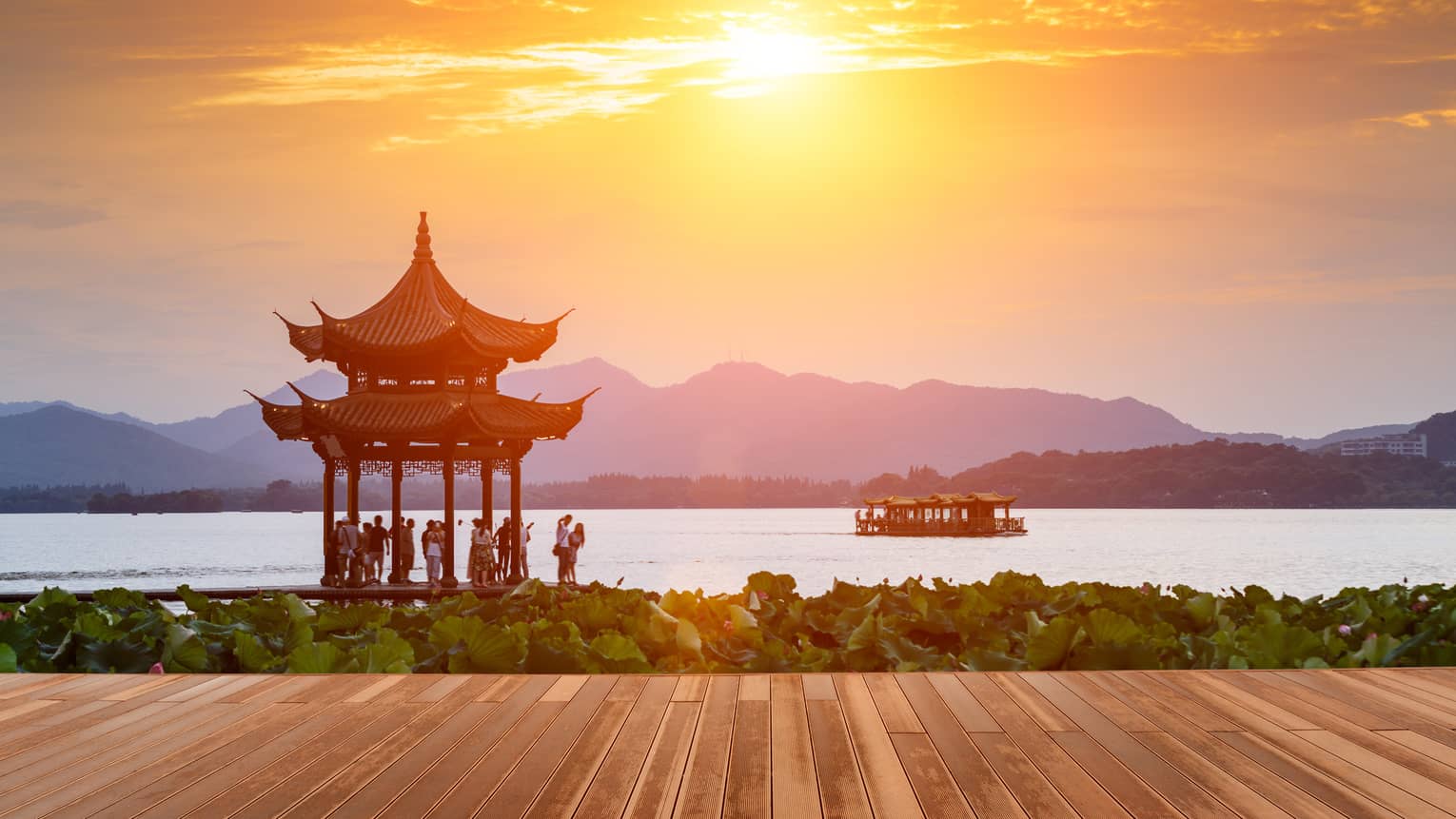 People gathered under gazebo by West Lake at sunset, boat on water 