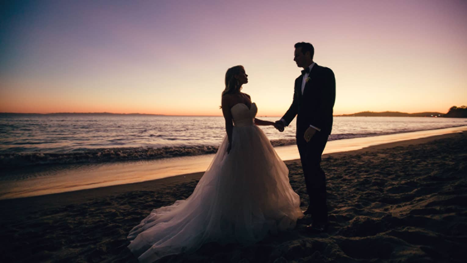 A silhouette of bride and groom standing by the ocean at dusk