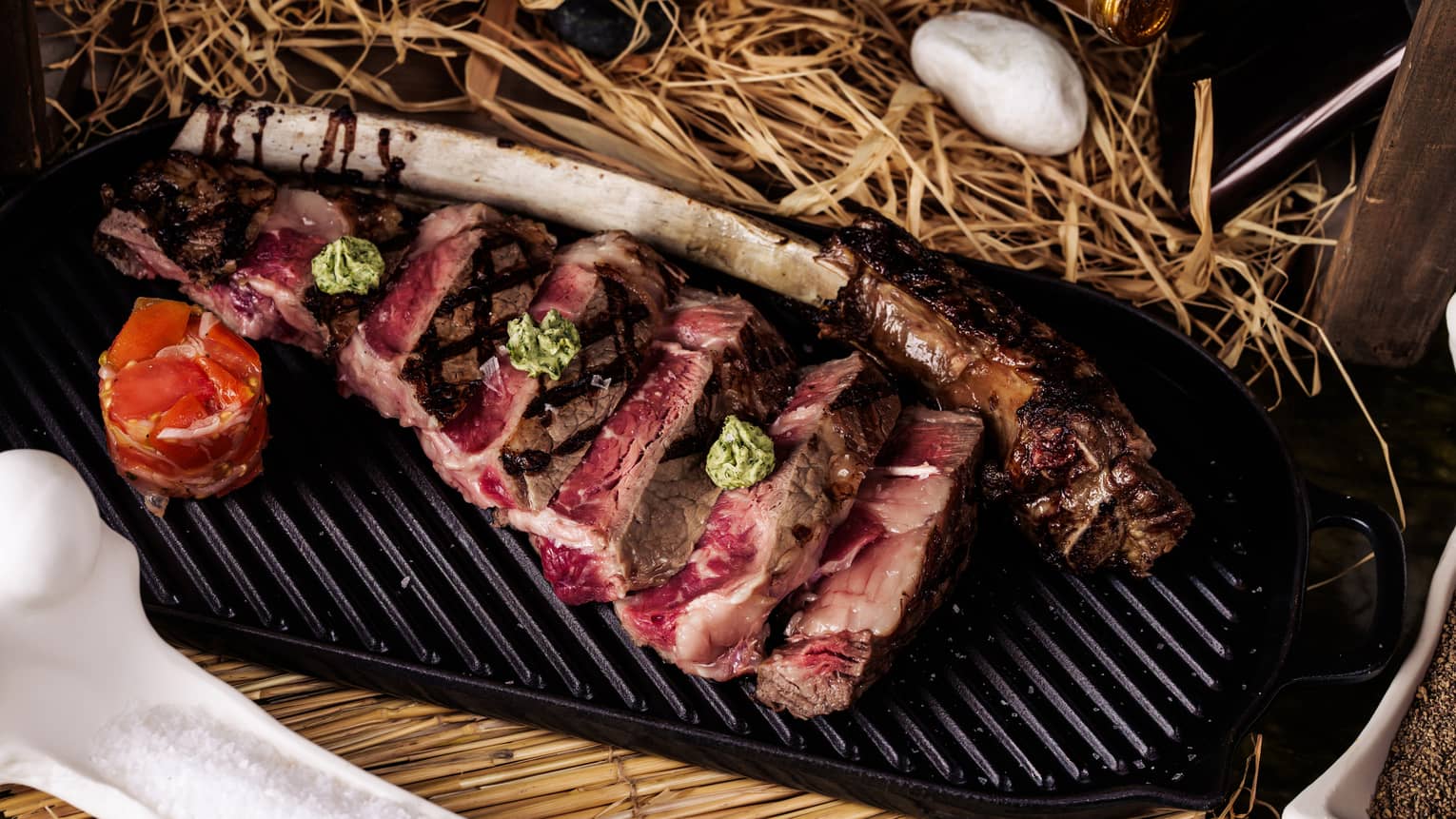 A sliced tomahawk steak with the bone next to it sits on a black griddle-style platter