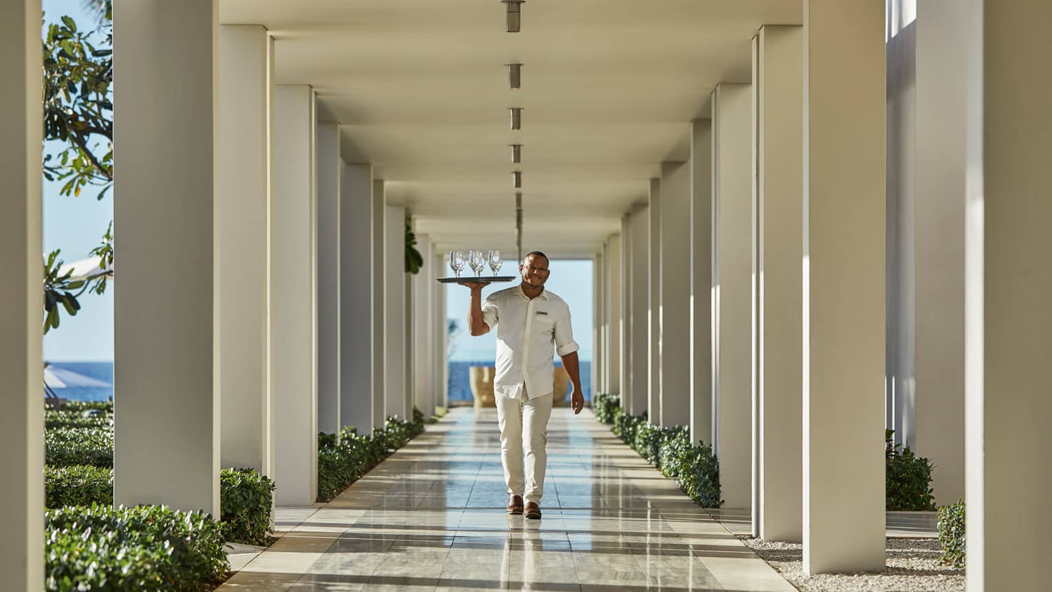 Server in white uniform carries tray of empty wine glasses through outdoor hallways with white posts, beams