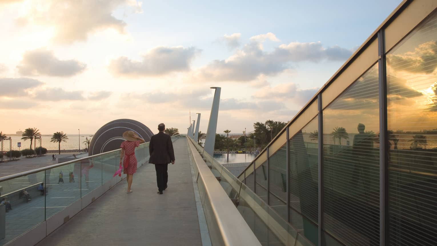 Back view of man in suit and woman in dress walking along raised Bibliotheca Alexandrina walkway at sunset