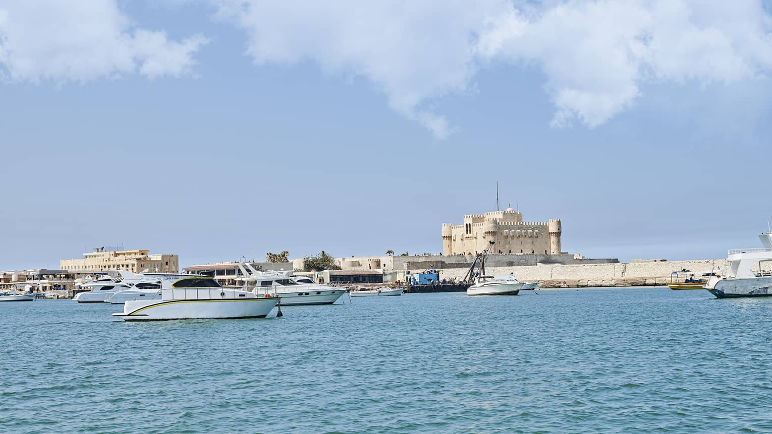 A distant sand-coloured fortress perched on a quay along a shore dotted with small yachts and fishing boats over calm waters.