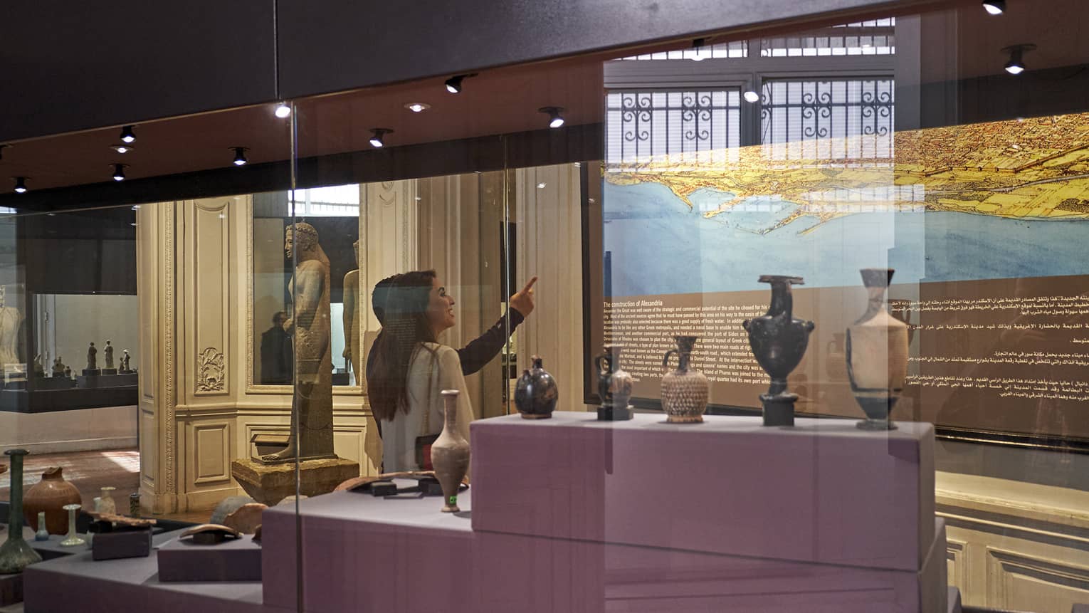 A visitor in a museum points up at a historical map lit up on a wall, behind them a display case with various vases and jugs.