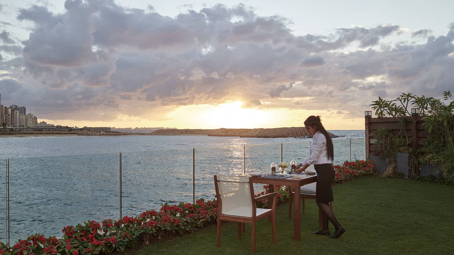 Woman sets a small wooden dining table on lawn next to sea