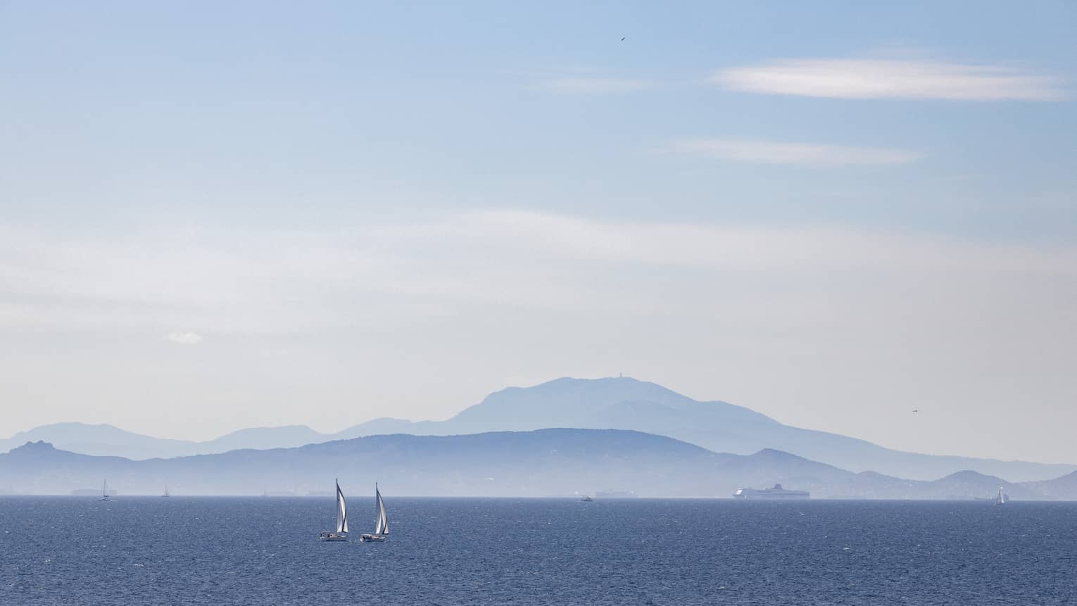 Two sailboats on blue ocean with blue-grey mountain range in the background 