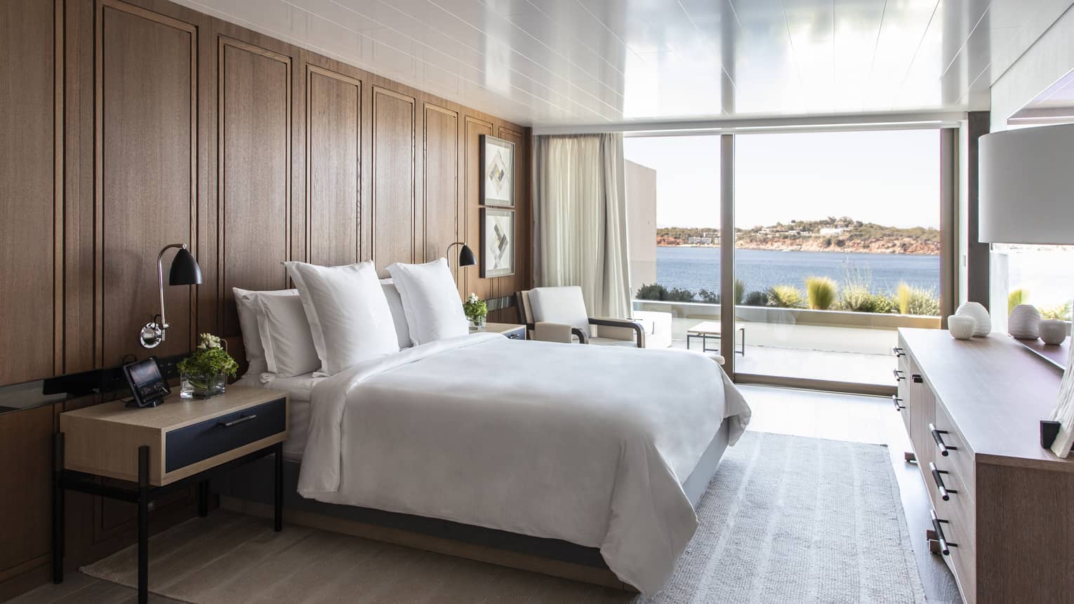 White king bed, wooden accent wall, contemporary night stand and dresser, wall of windows with sea view