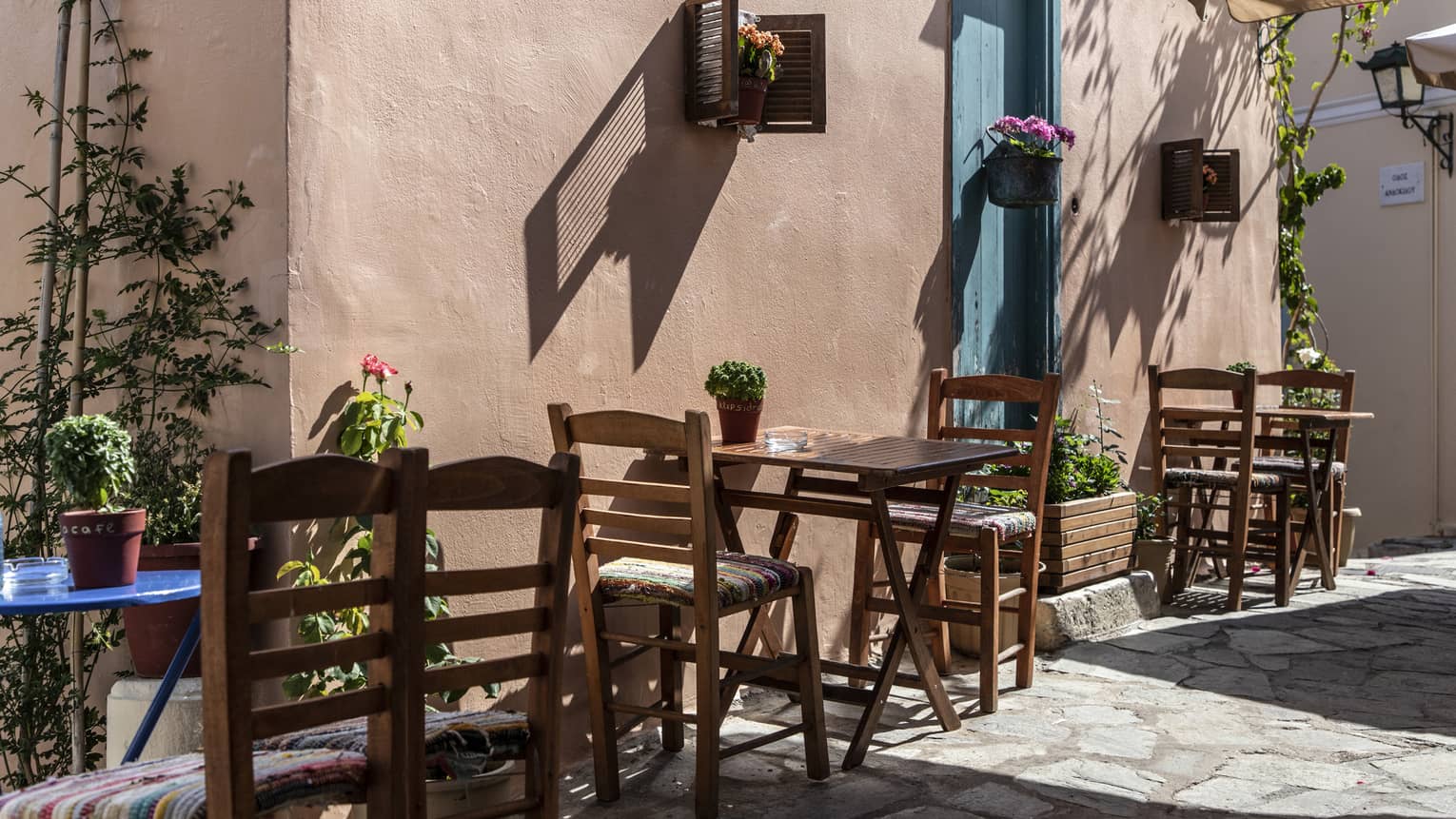 In terracotta-walled alley adorned with flowers and vines, wooden tables and chairs cast shadows on the cobblestone street.  