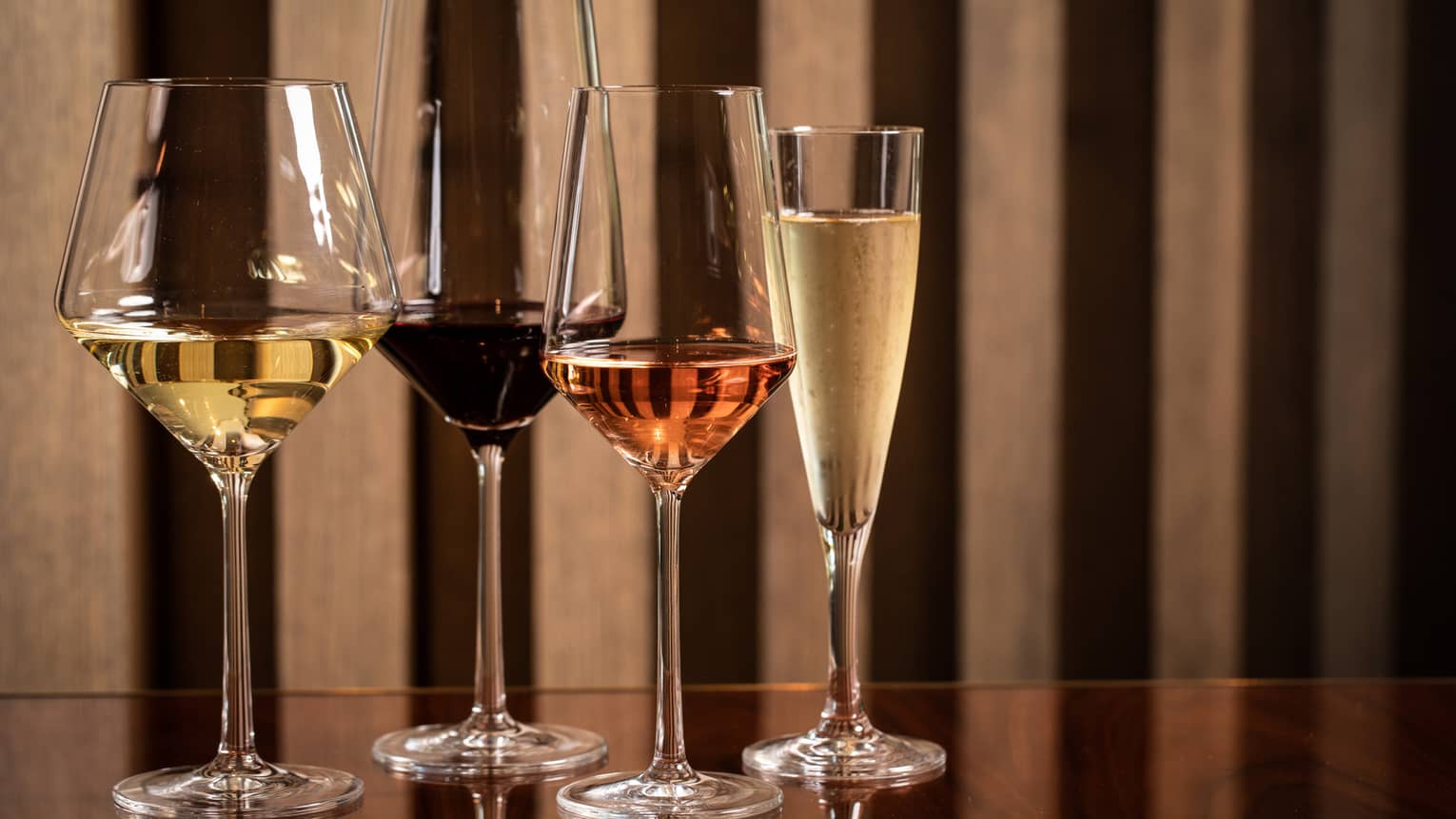 Four modern wine glasses holding white, red and rose wine are lined up on a wooden table 