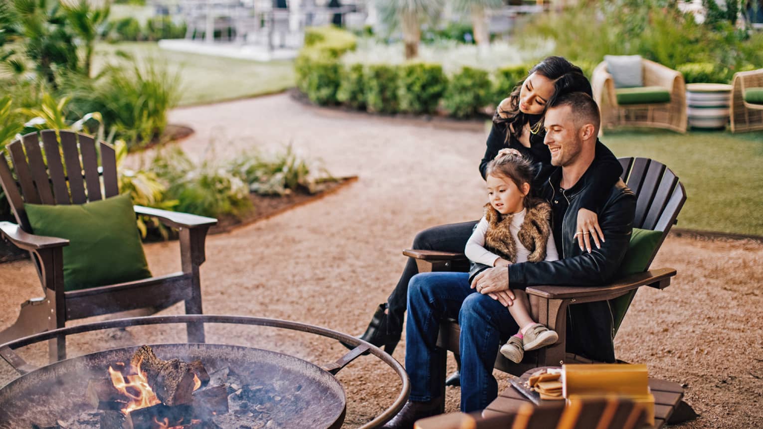 A family sitting on a wooden chair outside near a fire pit.
