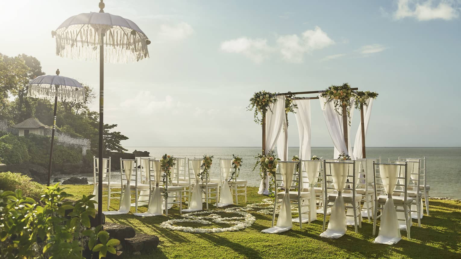 Sun shines over wedding altar draped in white fabric, rows of chairs overlooking ocean