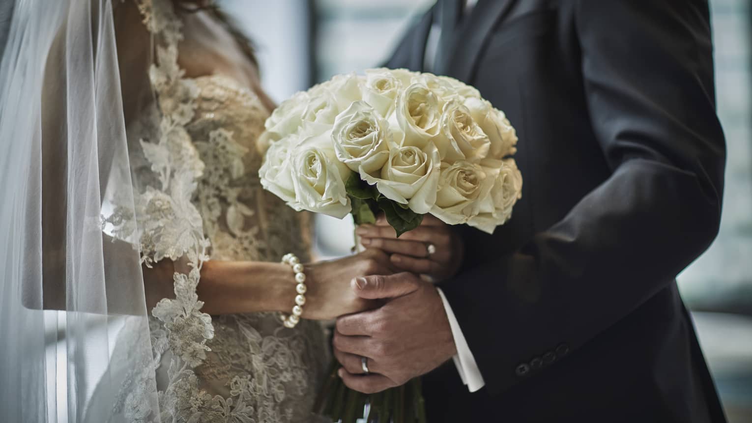 A close up of a bride in a lace gown and a groom in a gray suit hold the bride's bouquet of white roses together.