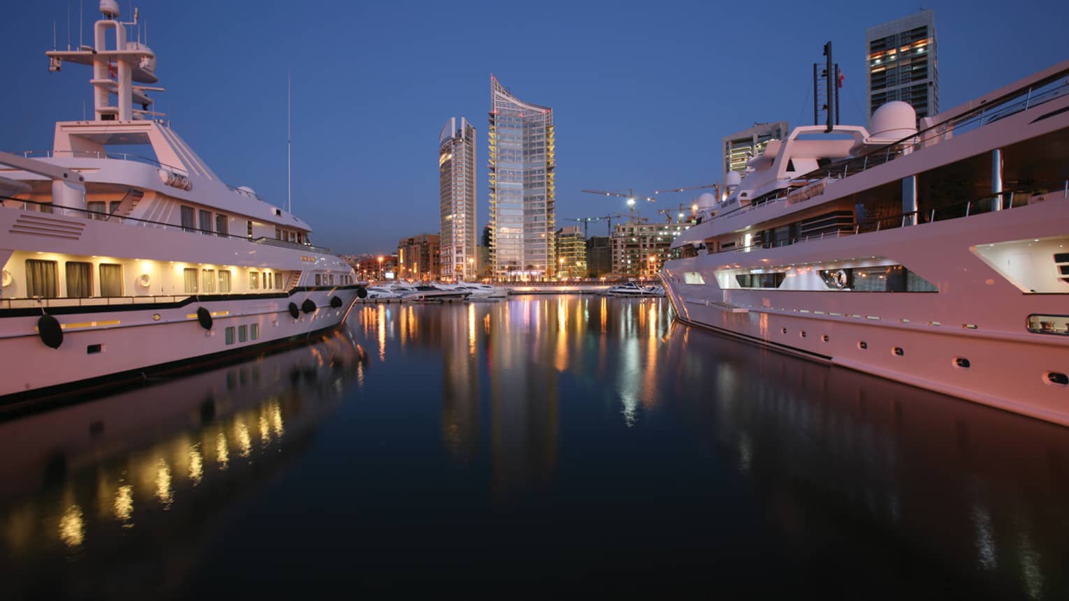 High rise buildings with lights at night reflected on water between two white yachts