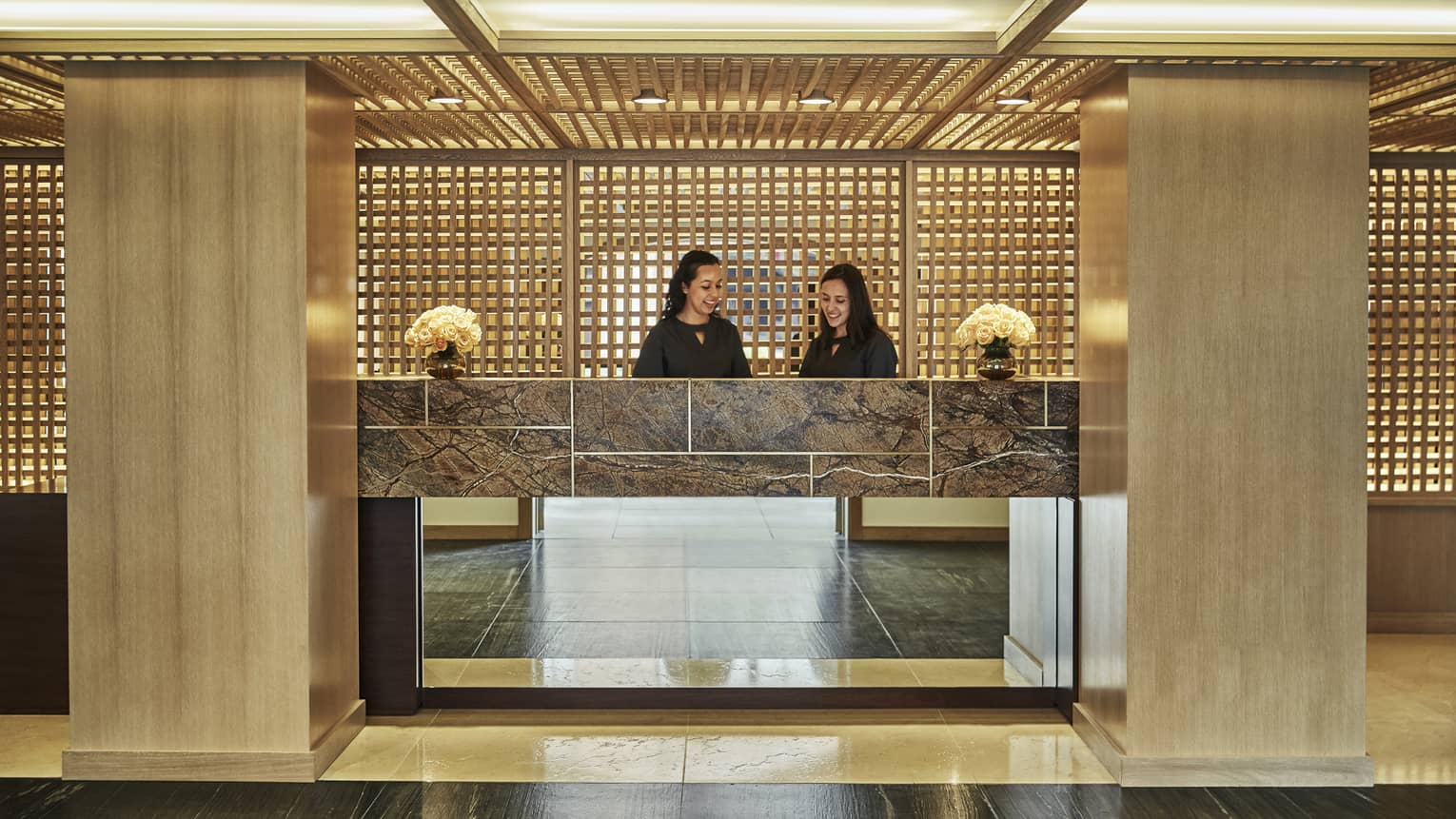 Two smiling hotel staff behind lobby front desk, wood pillars at side, wood lattice walls