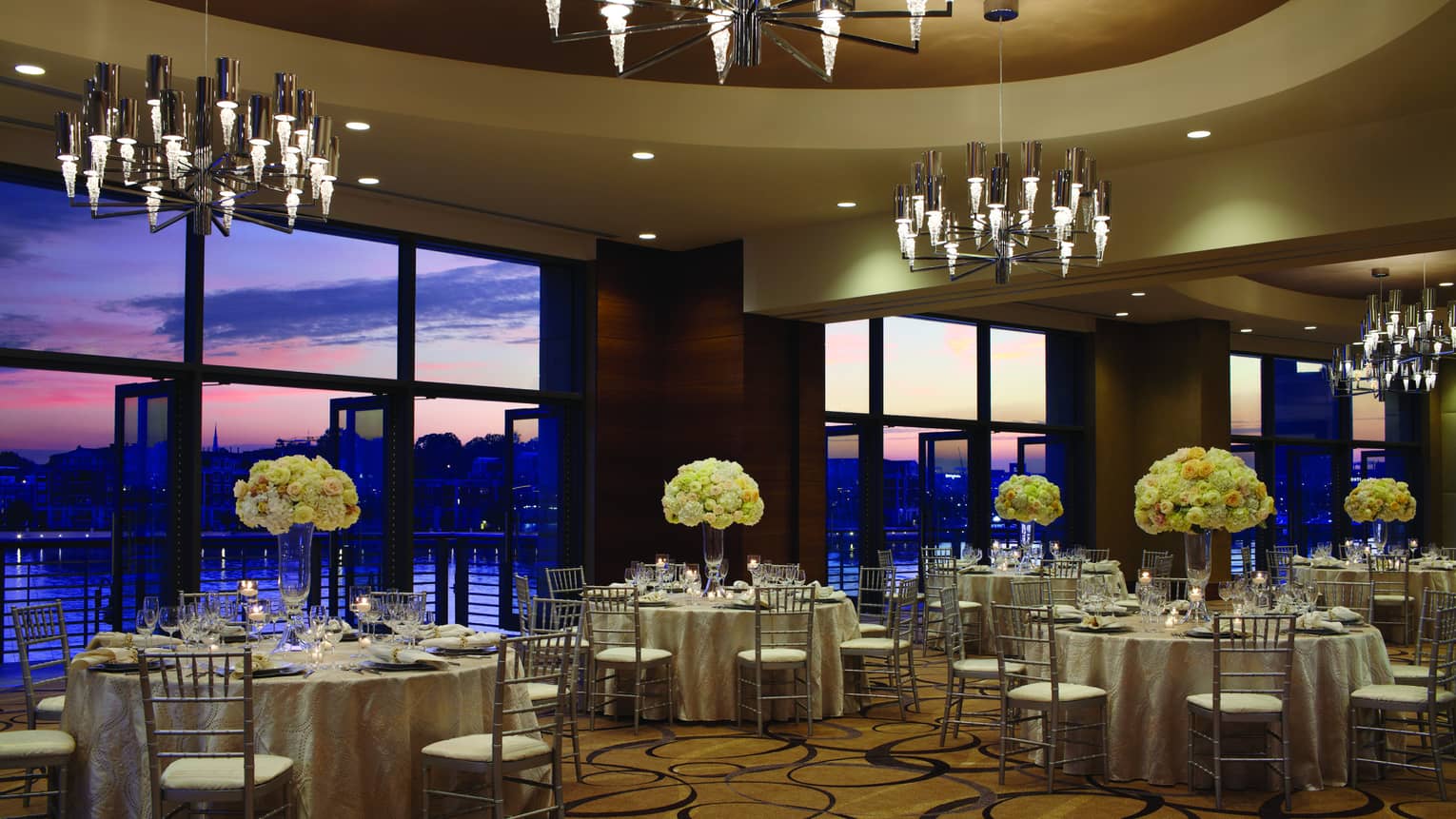Ballroom banquet dining tables under chandeliers and large windows with sunset Harbor views 