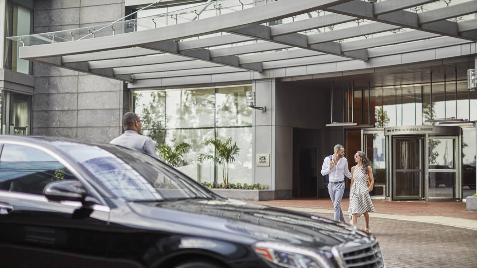 A man and woman walking towards a black sedan after leaving a hotel, with a hotel staff member standing by the car.