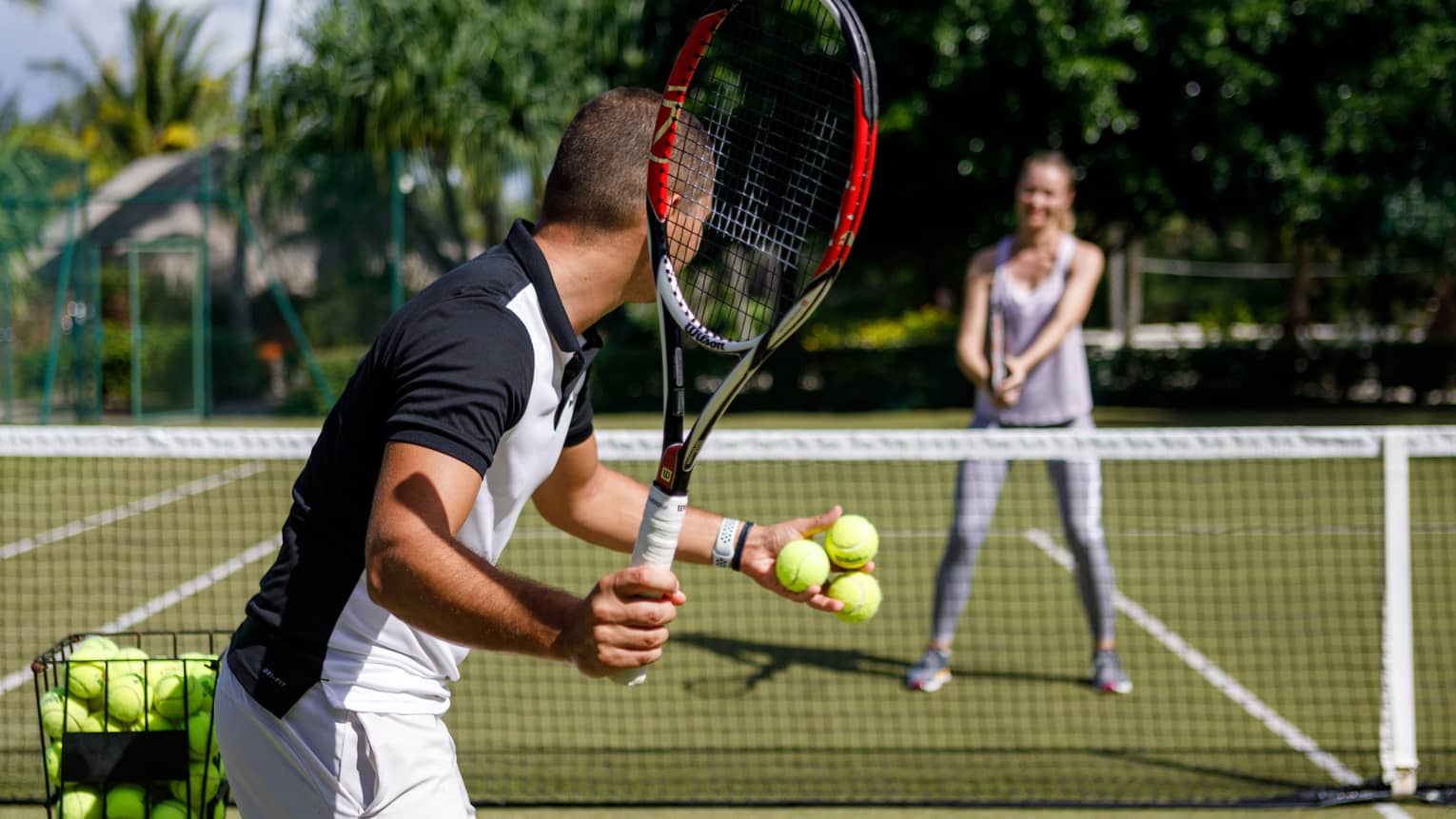 Couple playing tennis on court, man prepares to serve tennis balls to woman ready to swing