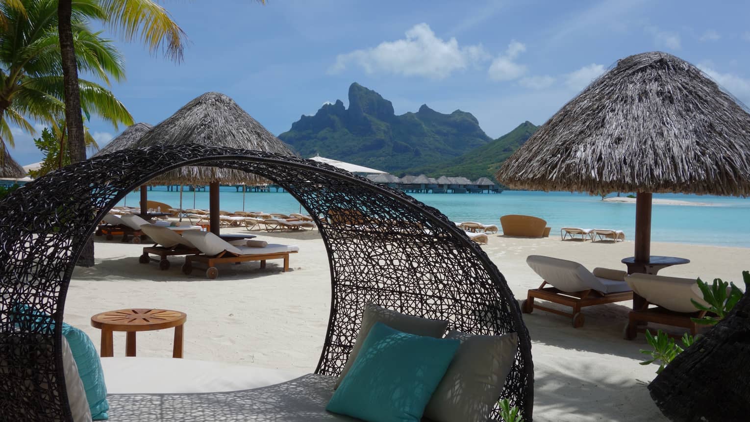 Large wicker patio bed, lounge chairs on sunny white sand beach