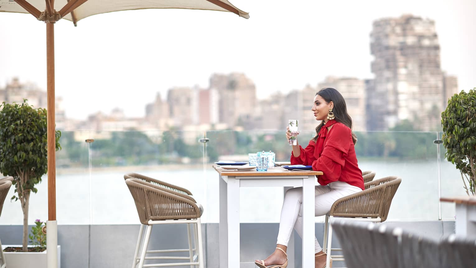 Closeup of woman in red shirt seated at table on Zoe terrace, holding a drink, Nile and city views