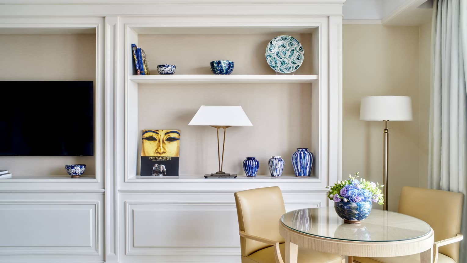 Small round table and two tan leather chairs set next to built-in wall shelves and TV