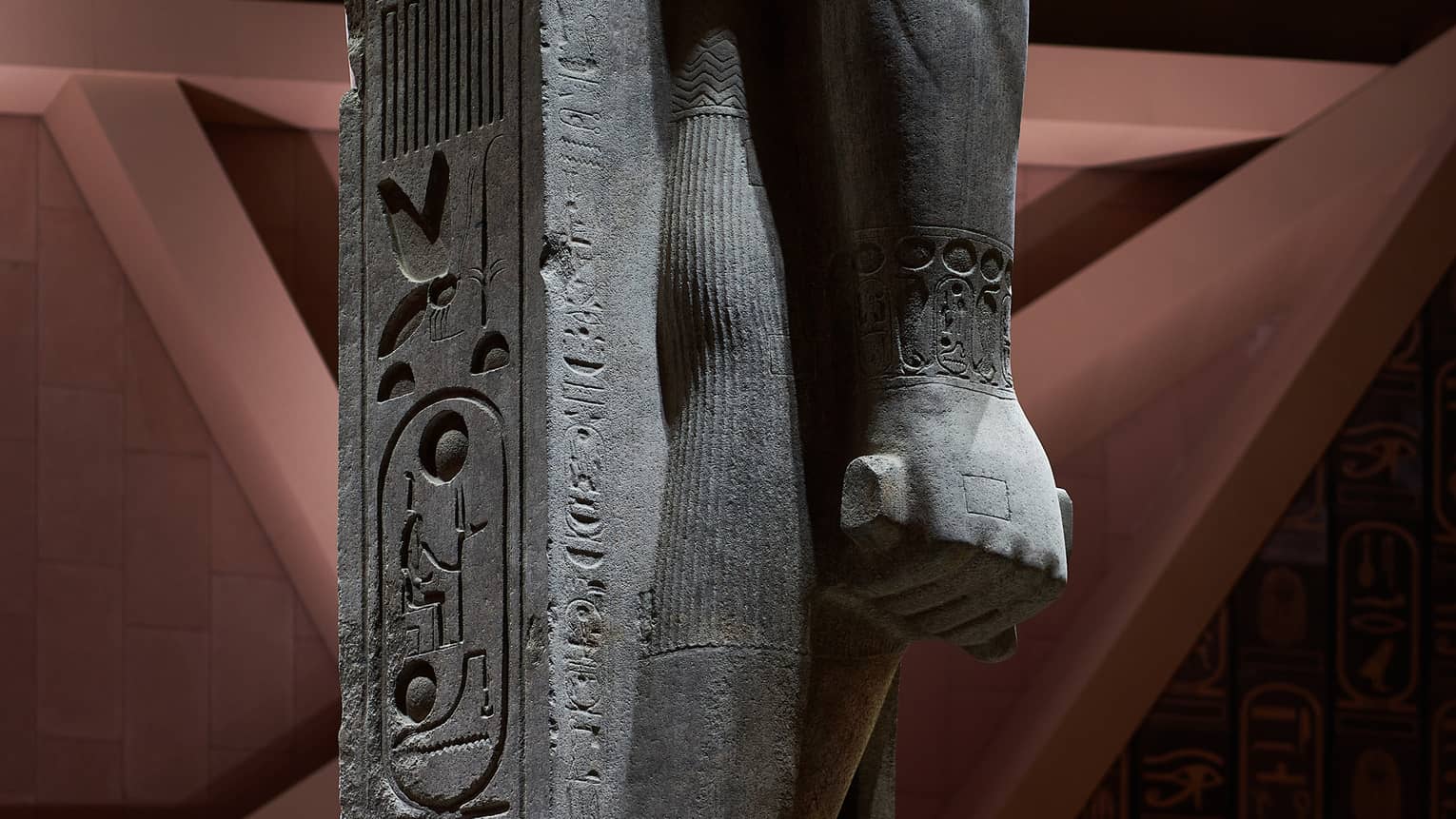 Close-up view of a statue's hand in a fist, supported by a stone inscribed with hyroglyph carvings. 