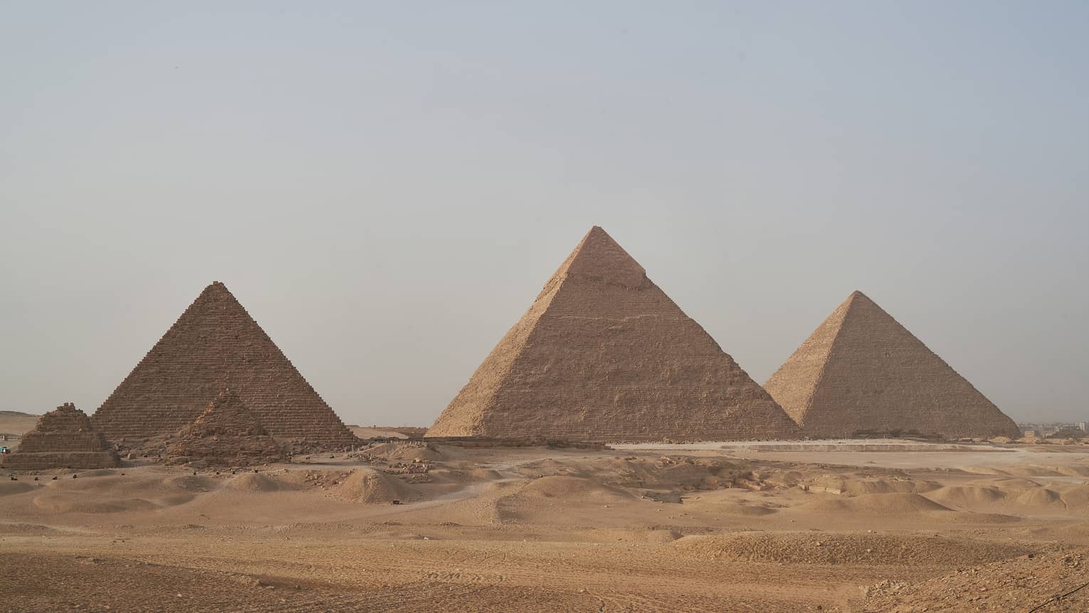 In the arid desert, a line of three towering pyramids dwarf two smaller pyramids before them, under a hazy blue sky. 