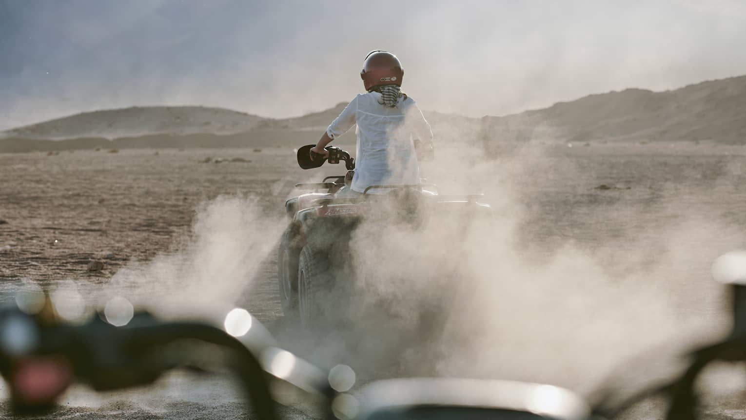 Rear view of an adventurer donning a red helmet while riding an ATV amid dust clouds; desert mountains ahead in the distance.