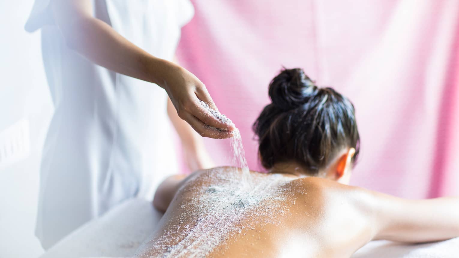 Spa staff pours salts, sand over woman's bare back as she lies on treatment room table