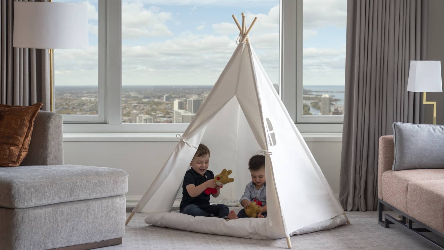 Two young boys playing in a small white tent in a hotel room, a large window looks out at a city behind them.