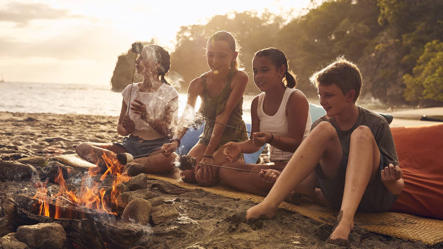 A group of young kids on a beach sitting near a small fire.