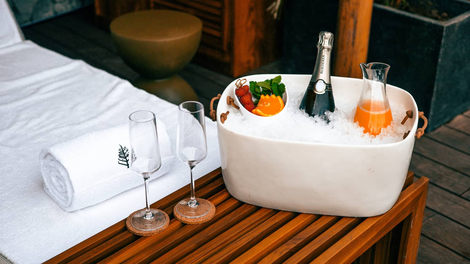 A white ceramic ice bucket holds a bottle of champagne, a glass carafe filled with fresh-squeezed orange juice and a small bowl of fruit, set next to two champagne flutes on a slated wooden bench