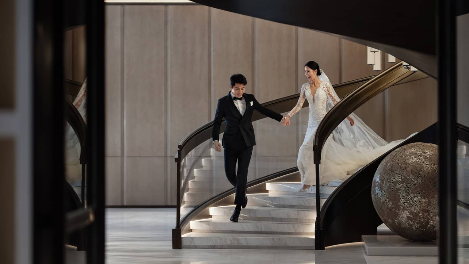 A smiling bride and groom walk hand in hand down the stairs at Four Seasons Hotel Dalian.
