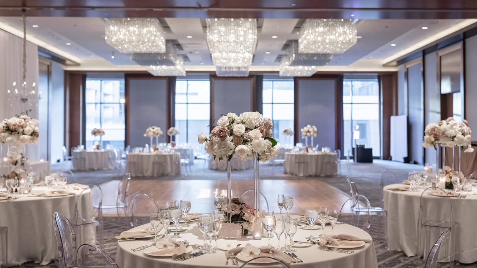 A well lit ballroom with ground tables and flower centerpieces and a section of hardwood floor in the middle.