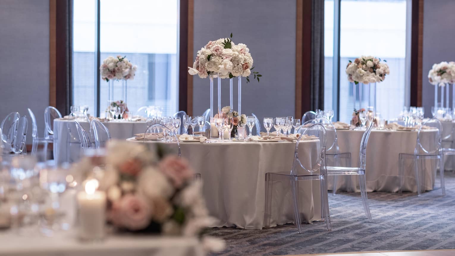 A group of round tables covered in cloth with flower centerpieces and clear chairs, large windows are behind the tables.