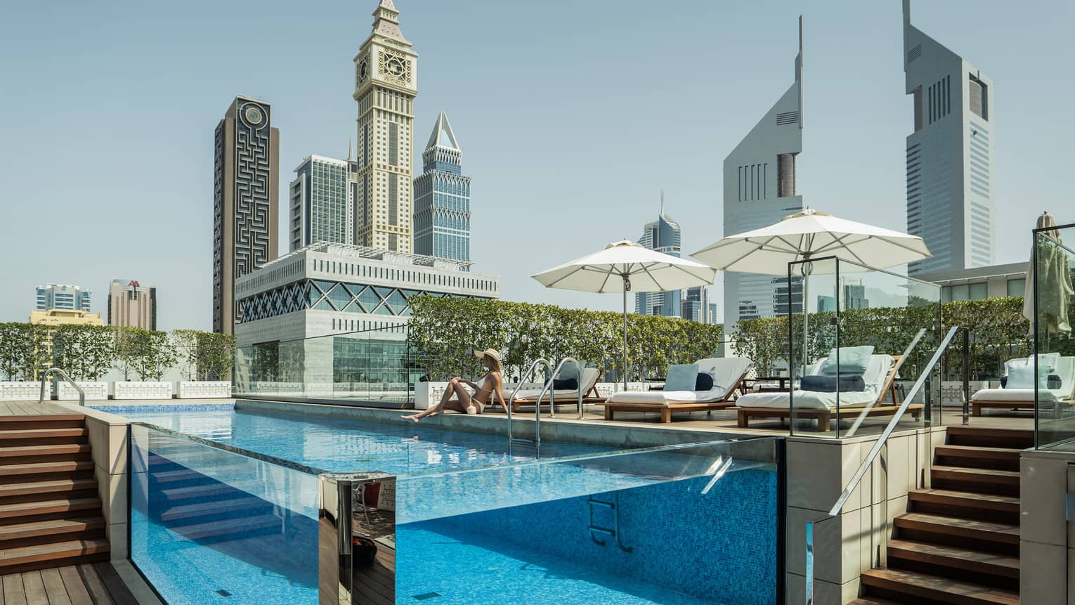 A guest lounges by a transparent rectangular rooftop pool, modern towers with geometric designs rising into the azure sky.