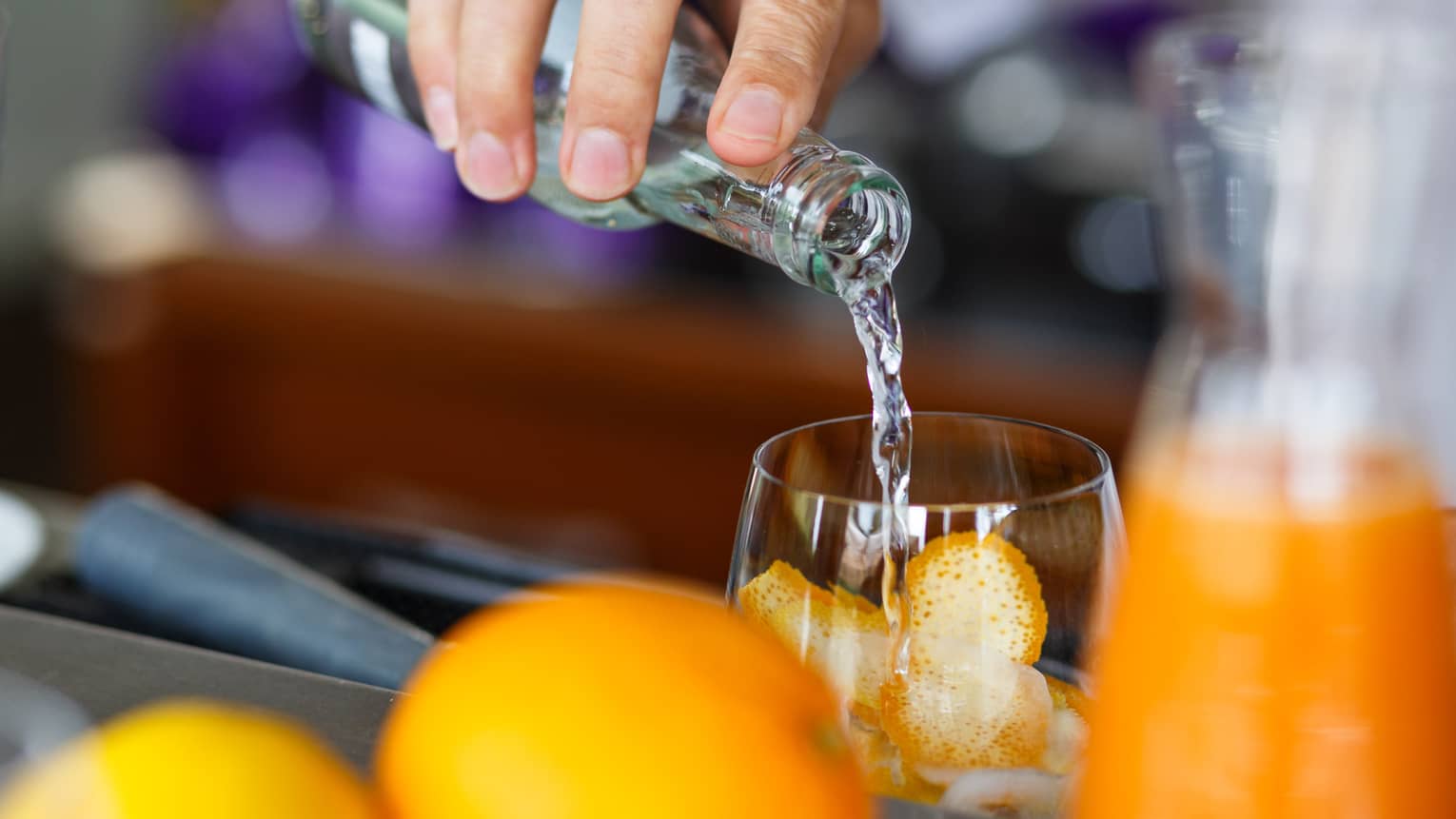 A hand pours clear liquid from a bottle into a glass filled with orange rind; in the foreground, whole oranges and a carafe.