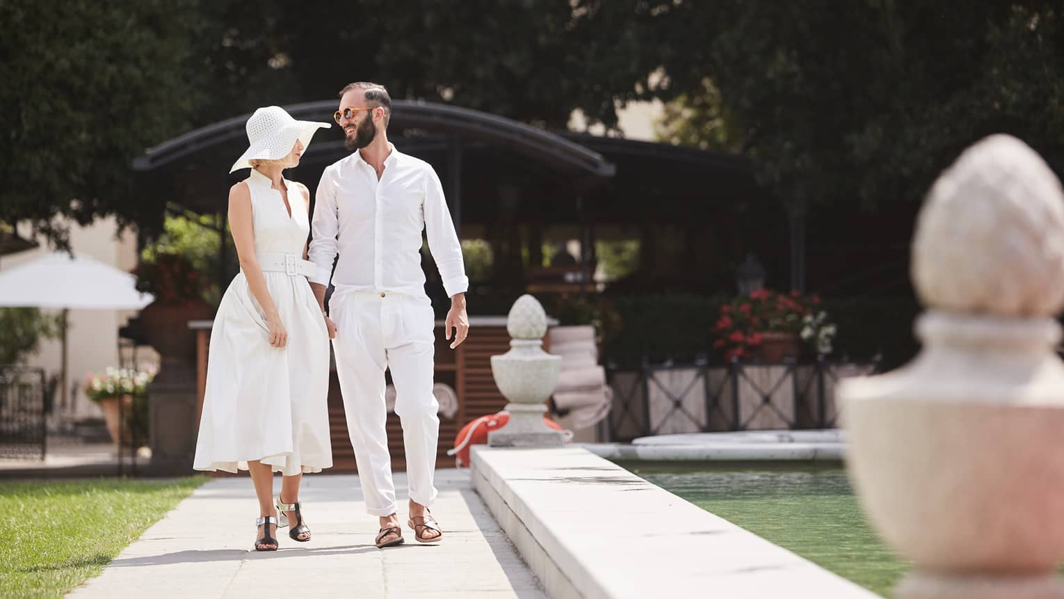 Man, woman wearing casual white cotton clothing stroll along sunny garden path by fountain