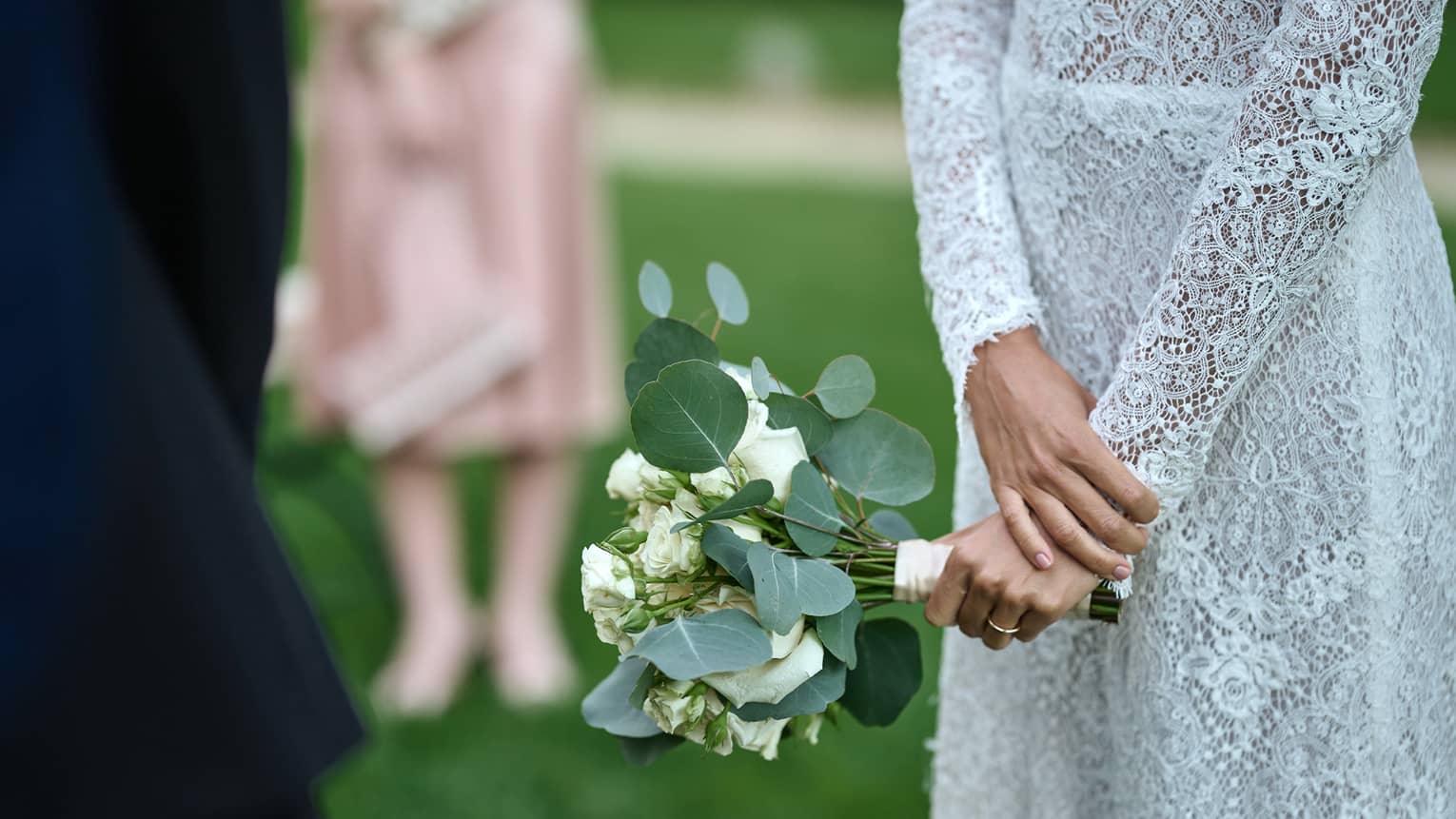 Torso of woman in simple lace wedding gown with hands together holding eucalyptus and white rose bouquet