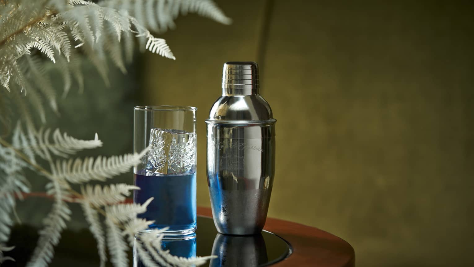 Blue cocktail in crystal highball glass beside chrome cocktail shaker and fern