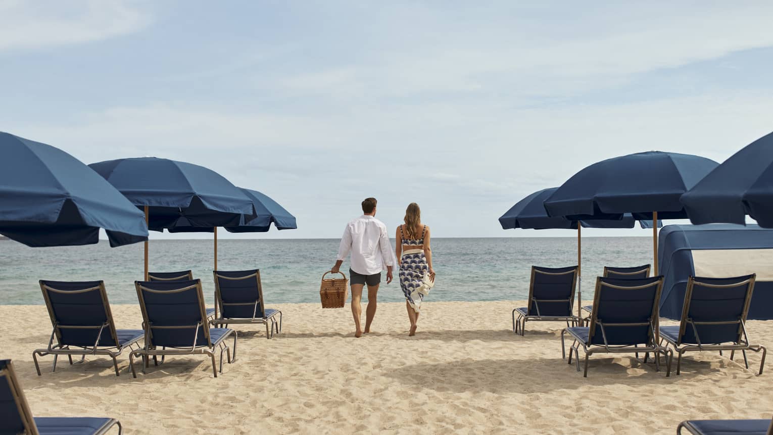 A man and woman walking between blue lounge chairs and umbrellas on sand towards the ocean.