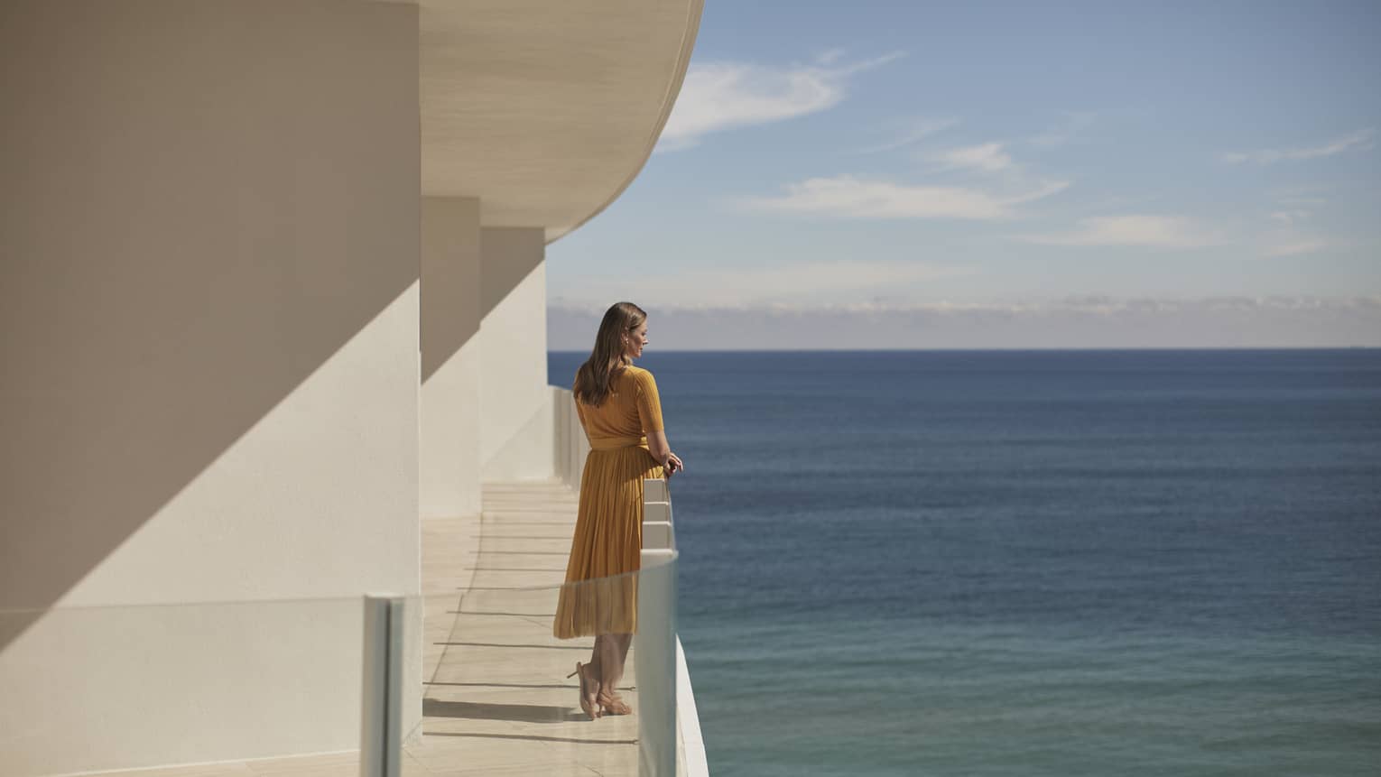 A woman standing on a balcony  of a white stone building with the ocean in the background.