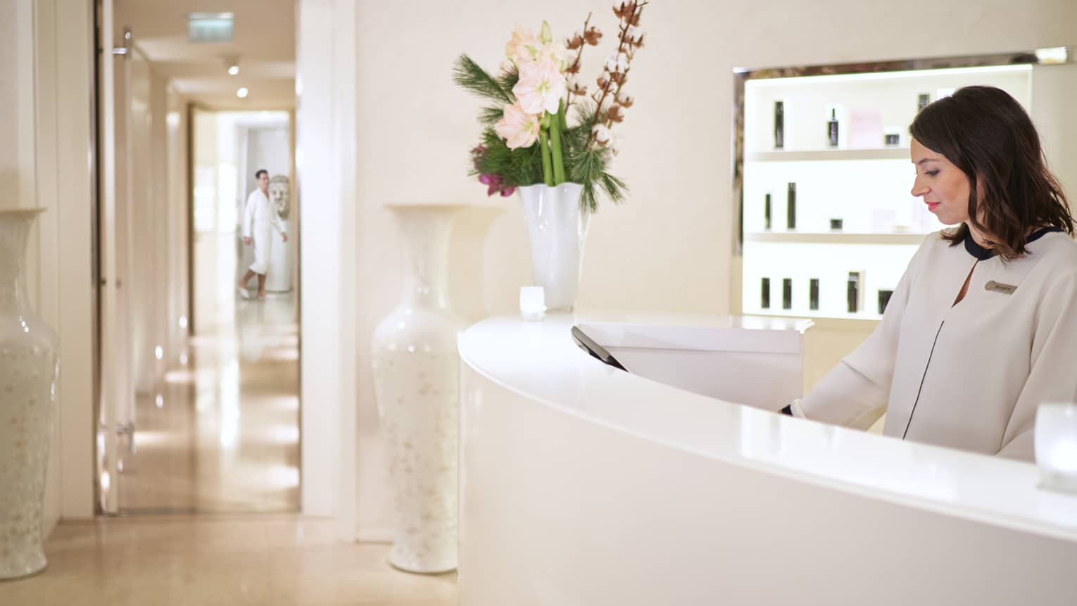 Staff member in white jacket standing behind curved white reception desk with flower vase, view into spa hallway
